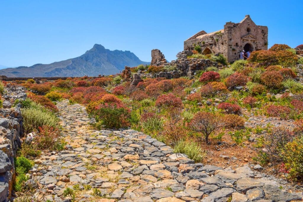 Bright red and yellow shrubs line a stone path leading to the ruins of a church at the venetian fortress on Gramvousa island Crete