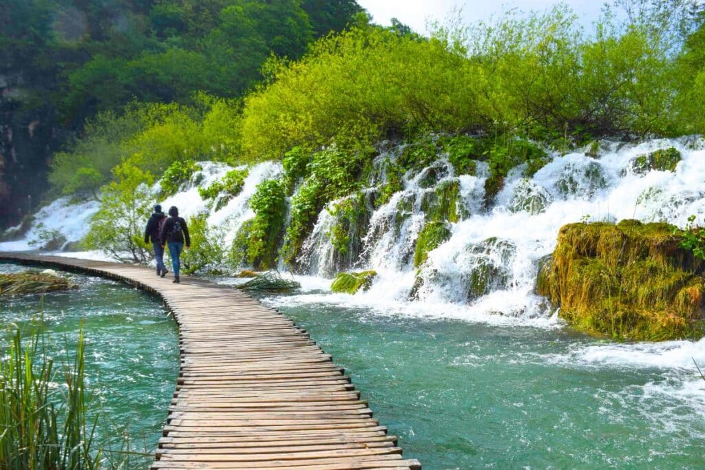 Visitors walk along a wooden pathway at Plitvice Lakes National Park in Croatia, surrounded by lush greenery and cascading waterfalls.