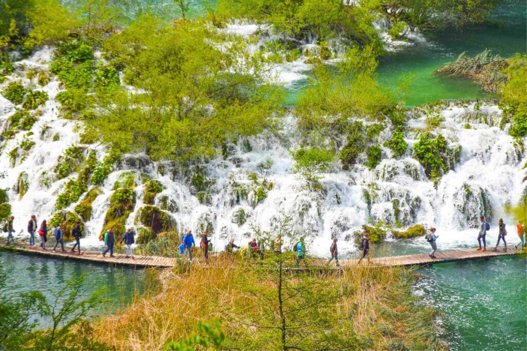 Visitors walk along a wooden pathway at Plitvice Lakes National Park in Croatia, surrounded by lush greenery and cascading waterfalls.