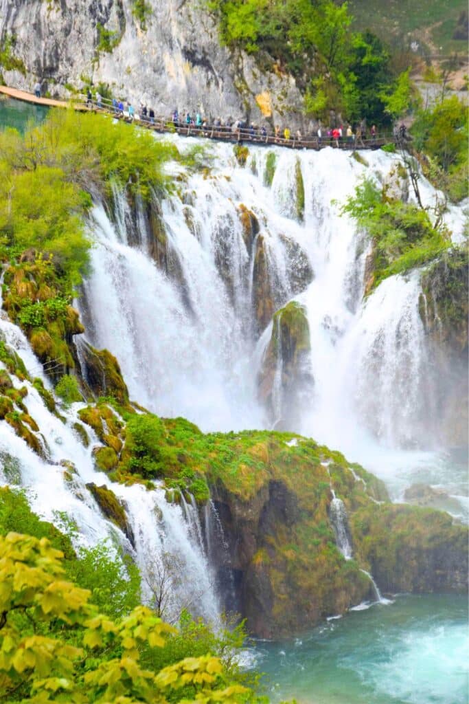 Visitors walk along a wooden pathway at Plitvice Lakes National Park in Croatia, surrounded by lush greenery and cascading waterfalls.